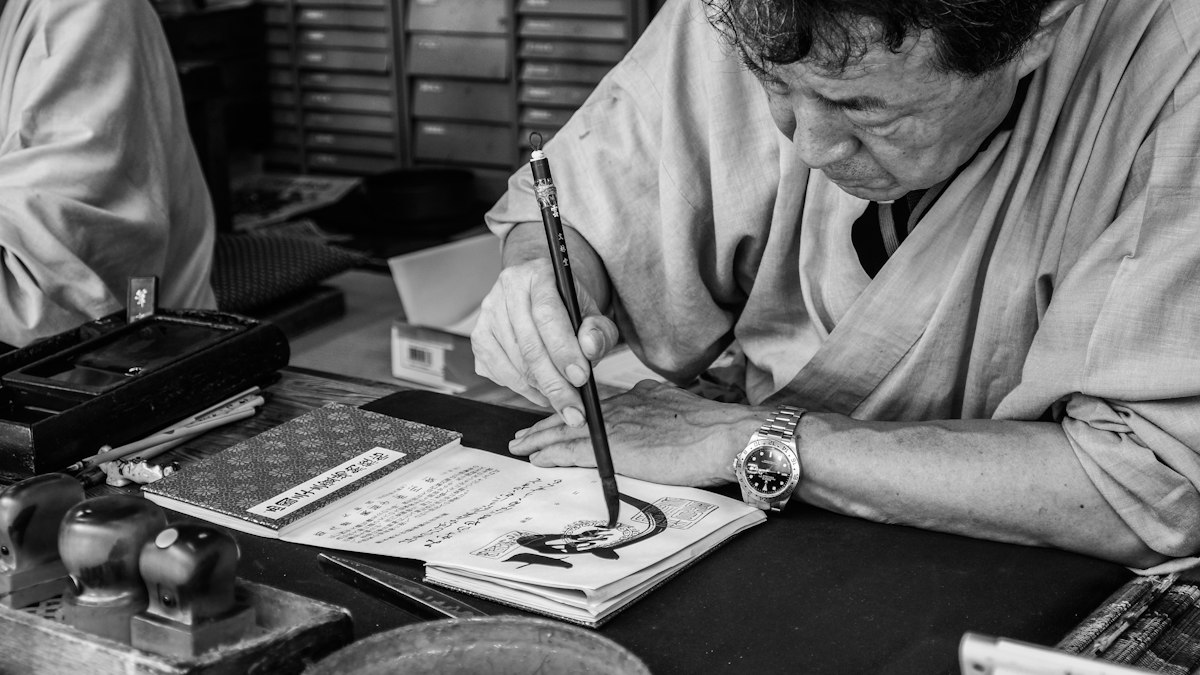 机に向かい筆で字を書く職人のモノクロ写真