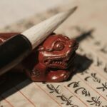 漢字が縦に並ぶ書の上に置かれた筆と朱色の獅子の筆置き